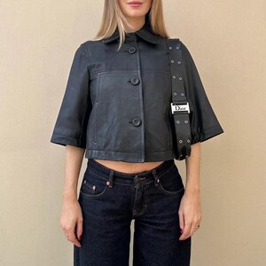 May include: Black leather jacket with short sleeves and a button-up front. The jacket is paired with dark wash denim jeans. A black shoulder bag with a silver buckle is worn over the shoulder.