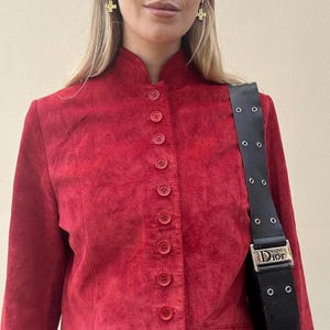 May include: A red suede jacket with a mandarin collar and a row of matching buttons down the front. The jacket has long sleeves and a tailored fit. A black shoulder bag with the word "Dior" is visible.