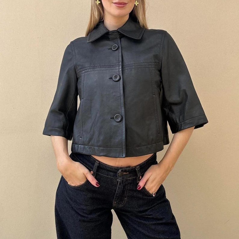May include: Black leather jacket with a collar and three buttons. The jacket has short sleeves and a cropped fit. The model is wearing dark wash denim jeans.