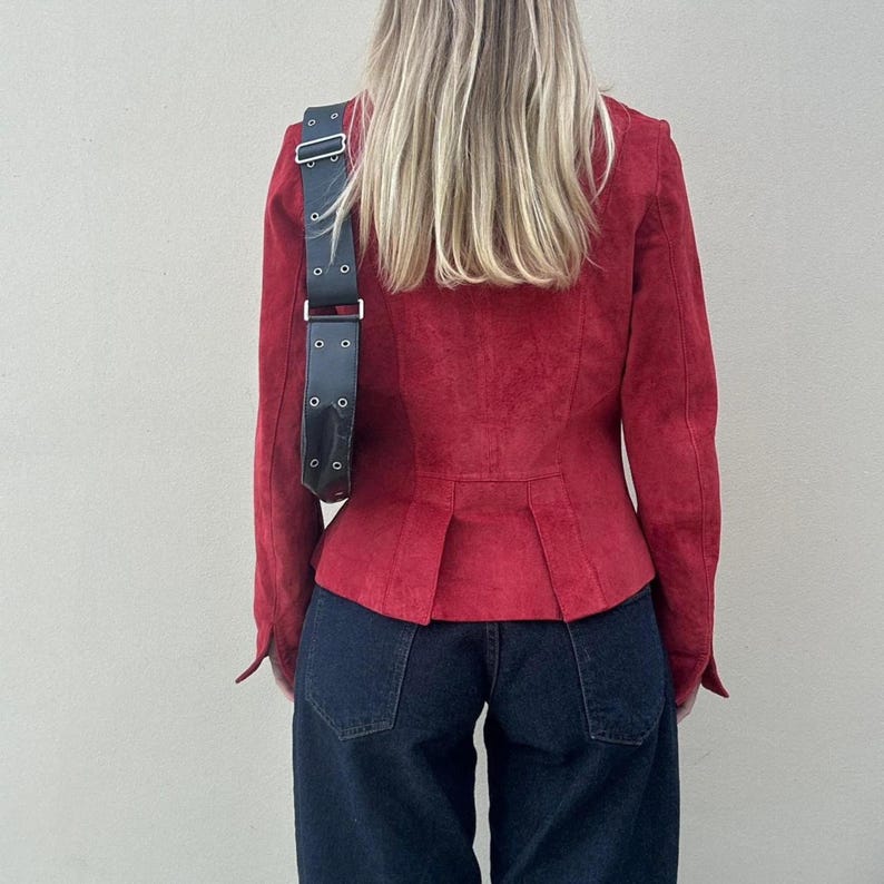 May include: A red suede jacket with long sleeves and a tailored fit. The jacket features a pleated back detail and is paired with dark wash denim jeans. A black shoulder bag with silver hardware is worn over the shoulder.
