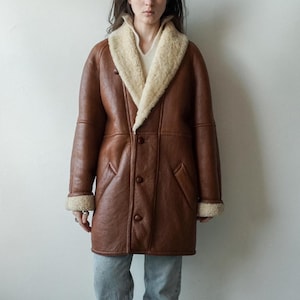 May include: A brown leather coat with a cream-colored shearling collar and cuffs. The coat has a button-down front, two pockets, and a classic, longline silhouette. The coat is worn over a cream-colored sweater and blue jeans.