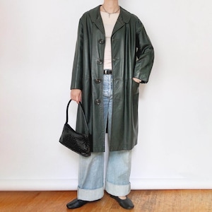 Op de afbeelding: Een lange, donkergroene leren jas met een knoopsluiting. De jas wordt gedragen met lichtblauwe jeans en zwarte ballerina's. Een zwarte handtas wordt in de hand gehouden. De jas heeft een klassieke, vintage stijl.