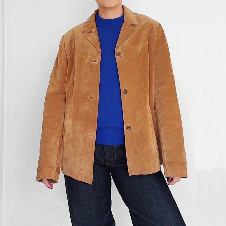 May include: A tan suede blazer with three buttons, a collared neckline, and long sleeves. The blazer is worn over a blue sweater and paired with dark blue jeans. The jacket has a classic, tailored design.
