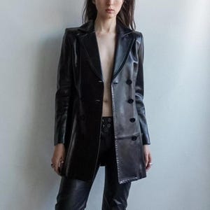 May include: A black leather blazer with a double-breasted design and long sleeves. The blazer has a notched lapel and button closures. The model is also wearing black leather pants. The outfit is a sleek, edgy style.