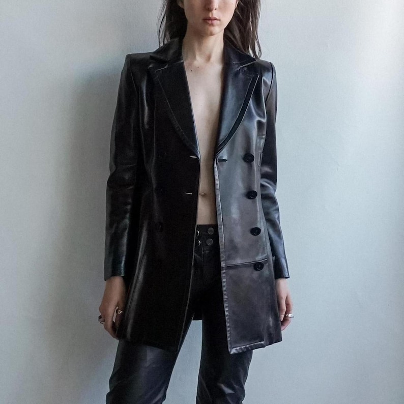 May include: A black leather blazer with a double-breasted design and long sleeves. The blazer has a notched lapel and button closures. The model is also wearing black leather pants. The outfit is a sleek, edgy style.