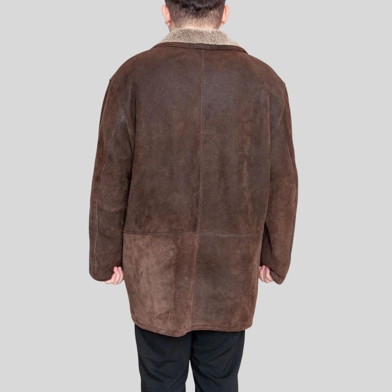 K&ouml;nnte beinhalten: Ein brauner Wildledermantel mit einem Shearling-Kragen. Der Mantel ist knielang und hat ein klassisches Design. Die R&uuml;ckansicht zeigt die Paneele und N&auml;hte des Mantels. Der Mantel wird &uuml;ber einer schwarzen Hose getragen.