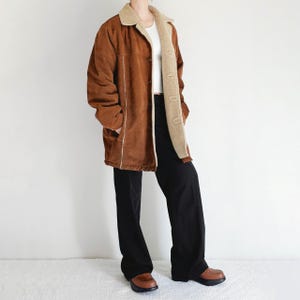May include: A brown suede coat with a cream-colored shearling lining and trim. The coat has a button-up front and two pockets. The person is wearing black pants and brown shoes.