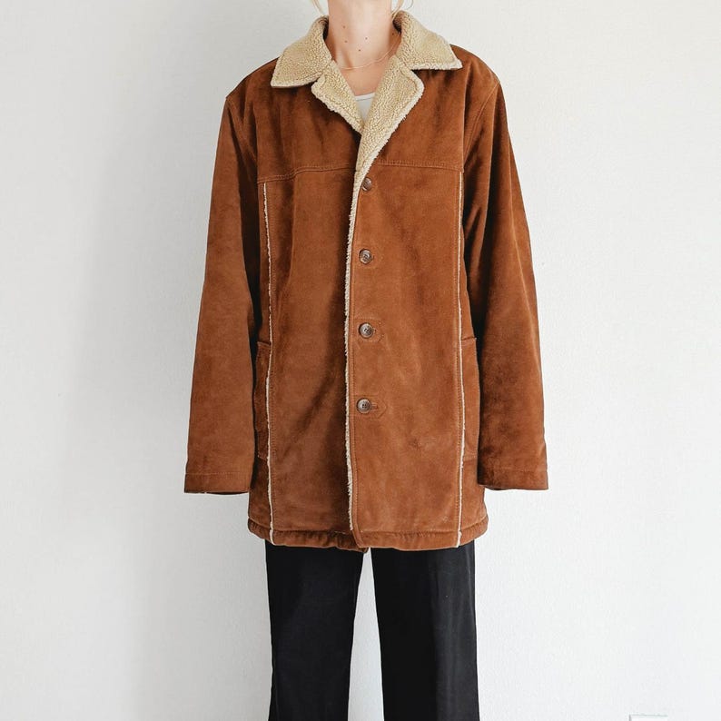 May include: A brown suede jacket with a cream-colored shearling collar and lining. The jacket has a button-up front, two front pockets, and cream-colored stitching along the seams. The jacket is worn over a white top and black pants.