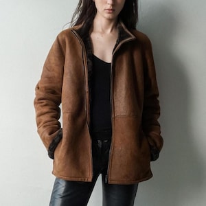 May include: A brown, zip-up jacket with a shearling lining. The jacket has a high collar and is worn over a black top and black pants. The jacket's exterior has a textured appearance.