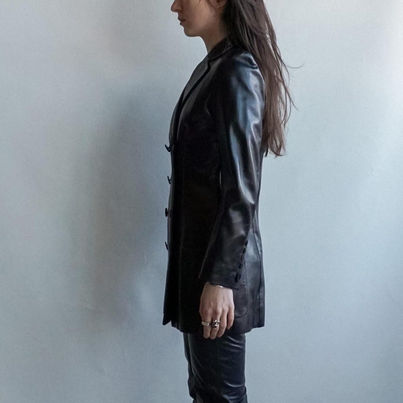 May include: A black leather blazer with a classic lapel collar and button closure. The longline jacket features long sleeves and a tailored fit. The blazer is paired with matching black leather pants. A stylish, timeless outerwear piece.