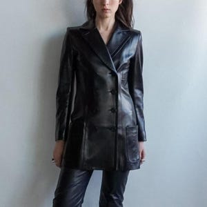 May include: A black leather blazer with a double-breasted design, featuring a notched collar, two front pockets, and button closures. The long-sleeved jacket is paired with black leather pants. The outfit is worn by a person.