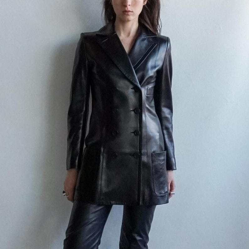 May include: A black leather blazer with a double-breasted design, featuring a notched collar, two front pockets, and button closures. The long-sleeved jacket is paired with black leather pants. The outfit is worn by a person.