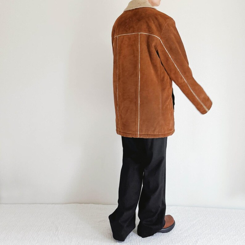 May include: A brown suede jacket with a cream-colored shearling collar and white stitching details. The jacket is worn with black pants and brown shoes. The jacket appears to be a medium length.