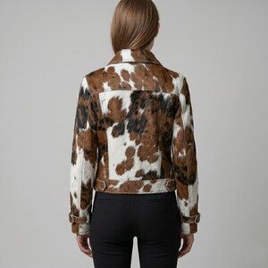 Puede incluir: Chaqueta de piel de vaca con un estampado de manchas marrones, negras y blancas. La chaqueta tiene un cuello cl&aacute;sico, pu&ntilde;os abotonados y un cintur&oacute;n en la cintura. La chaqueta se lleva sobre pantalones negros.