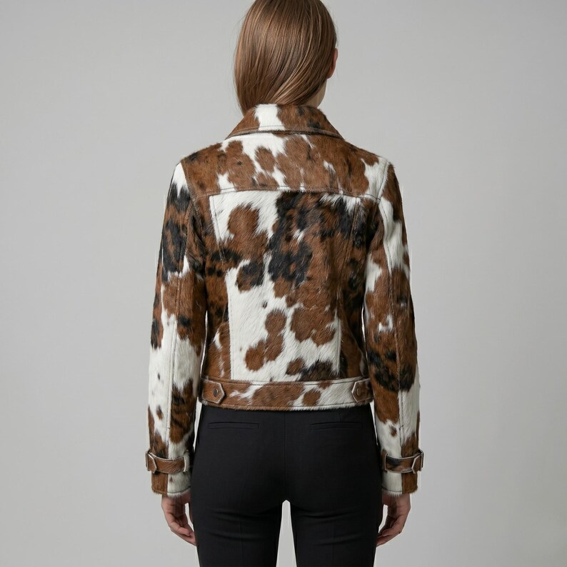 Puede incluir: Chaqueta de piel de vaca con un estampado de manchas marrones, negras y blancas. La chaqueta tiene un cuello cl&aacute;sico, pu&ntilde;os abotonados y un cintur&oacute;n en la cintura. La chaqueta se lleva sobre pantalones negros.