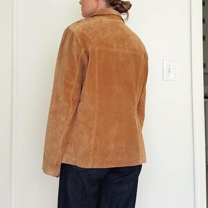 May include: A tan suede jacket with a collared neckline and button closure. The jacket has a classic design with a tailored fit. The garment is paired with dark blue denim jeans. The jacket is a neutral color, suitable for various outfits.