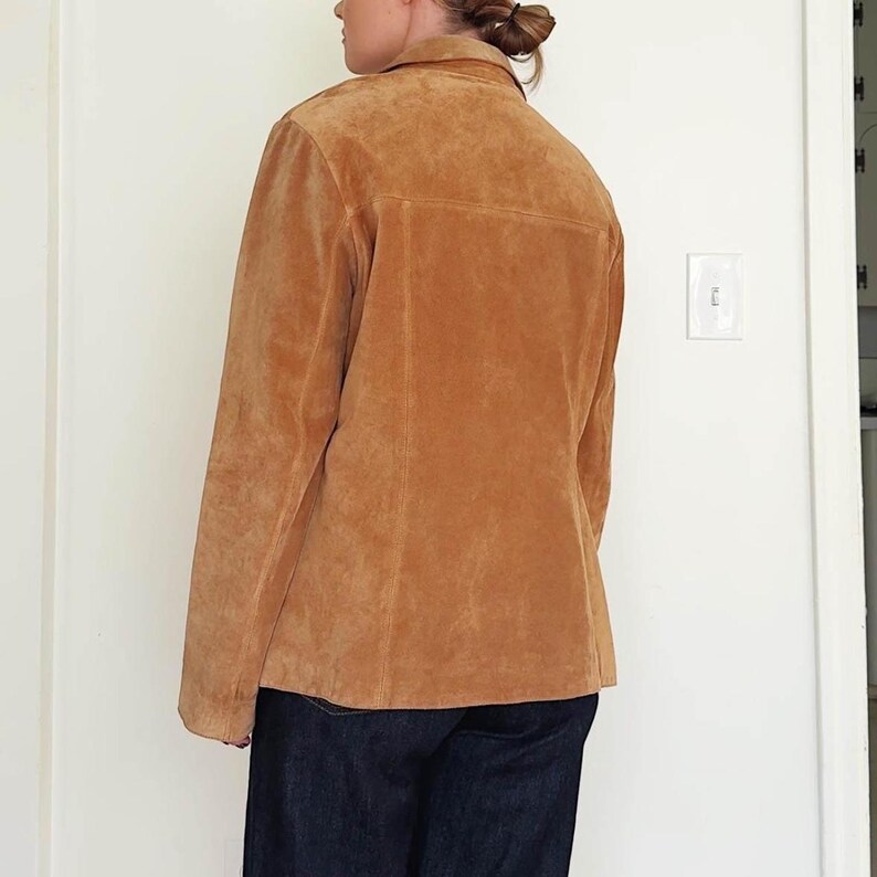 May include: A tan suede jacket with a collared neckline and button closure. The jacket has a classic design with a tailored fit. The garment is paired with dark blue denim jeans. The jacket is a neutral color, suitable for various outfits.