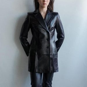May include: A black leather double-breasted blazer with a notched lapel and two front pockets. The blazer has a tailored fit and is worn with matching black leather pants. The jacket is approximately 36 inches long.
