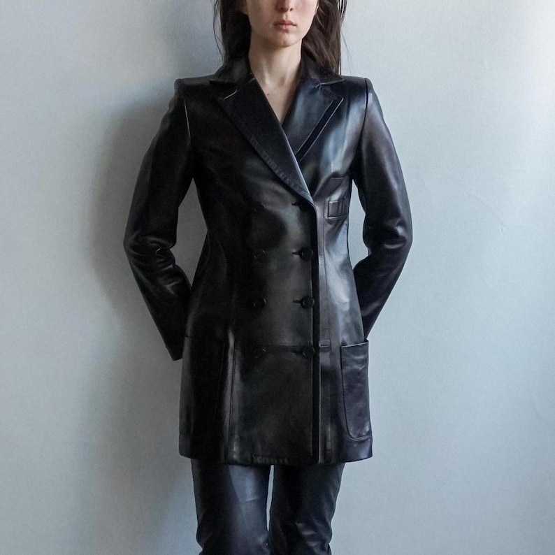 May include: A black leather double-breasted blazer with a notched lapel and two front pockets. The blazer has a tailored fit and is worn with matching black leather pants. The jacket is approximately 36 inches long.