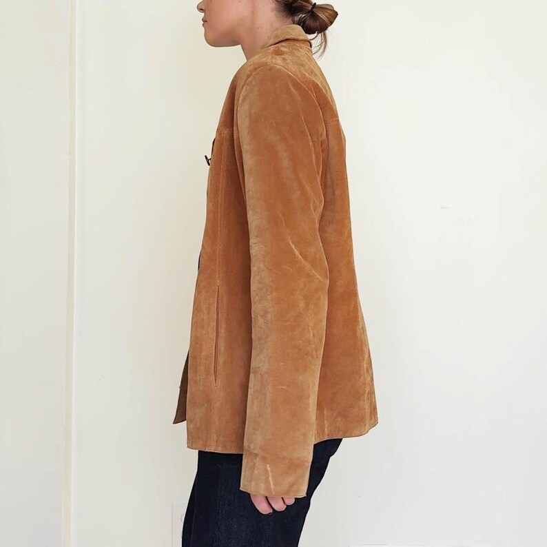 May include: A tan suede blazer with a classic collar and button closure. The jacket has long sleeves and a tailored fit. The blazer is worn with dark blue jeans. The background is a plain white wall.