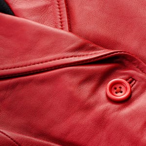 May include: Close-up of a red leather jacket, showcasing the texture and stitching details. A red button is visible, adding a pop of color. The jacket's design suggests a classic style, suitable for various occasions.