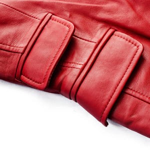 May include: Close-up of a section of a red leather jacket, showcasing the texture and stitching details. The image highlights the flap and strap closures, emphasizing the craftsmanship and design of the garment. The leather appears smooth and supple.