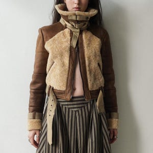 May include: A brown and tan shearling jacket with a high collar and a zip-up front. The jacket has brown leather sleeves and accents, with tan shearling panels on the chest and back. The jacket has a belted waist and cuffs. The model is wearing a striped skirt.