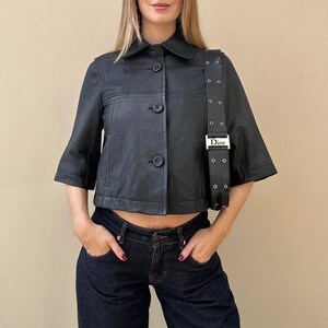 May include: Black leather jacket with a collar and three buttons. The jacket has short sleeves and is paired with dark wash denim jeans. A black shoulder bag with silver hardware is also visible.
