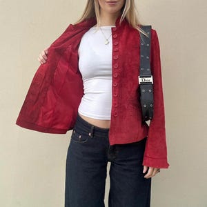 May include: A red suede jacket with a mandarin collar and button closure. The jacket is paired with a white top and dark wash denim jeans. A black shoulder bag with silver hardware is worn across the body.