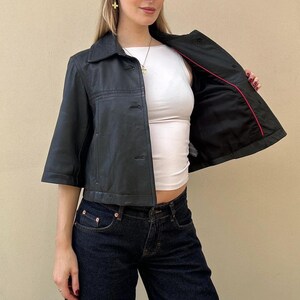 May include: A black leather jacket with short sleeves and a collared neckline. The jacket is buttoned and has a red lining. The model is wearing a white top and dark blue jeans.