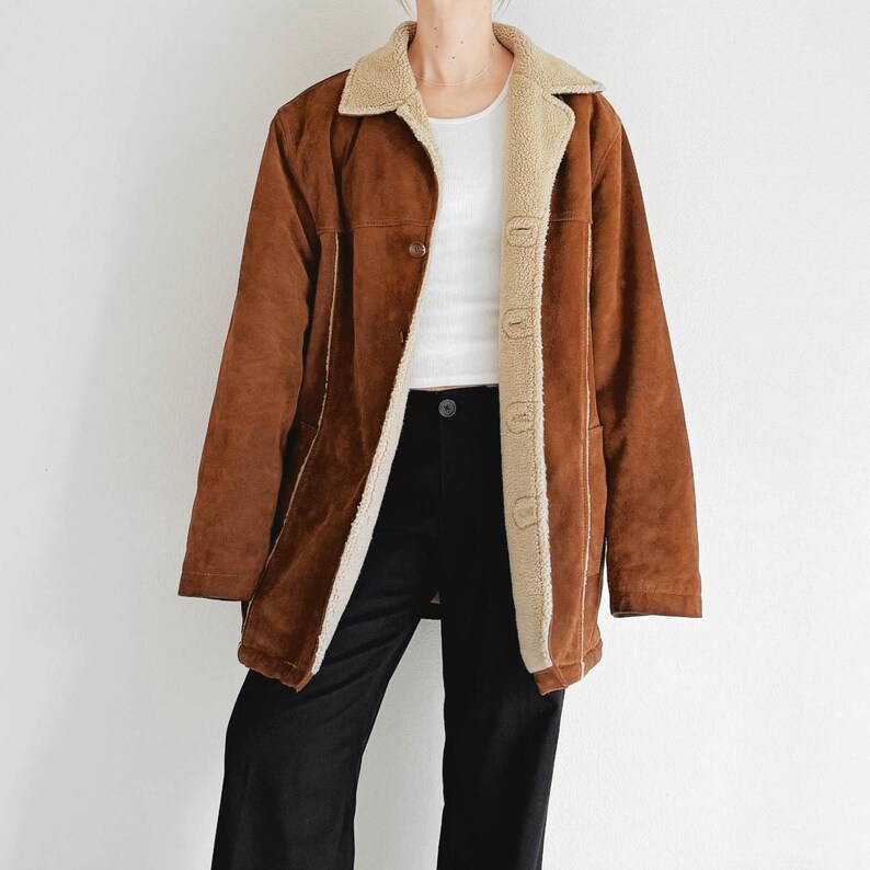 May include: A brown suede jacket with a cream-colored shearling lining and collar. The jacket has a button closure and cream-colored trim. The model is wearing a white top and black pants.