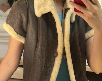 Könnte beinhalten: Eine braune Lederweste mit cremefarbenem Shearling-Futter und -Besatz. Die Weste hat kurze Ärmel und einen Knopfverschluss. Der Shearling-Besatz ist am Kragen, an der Vorderseite und an den Ärmelrändern sichtbar.
