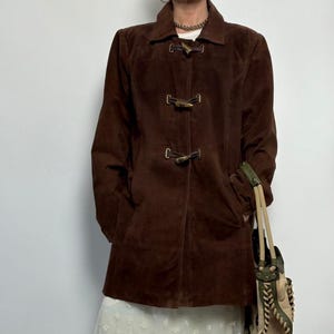May include: A brown suede coat with a collared neckline and wooden toggle closures. The coat has long sleeves and a tailored fit. A beige and green handbag with a braided strap is visible.