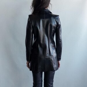 May include: A black leather blazer with a notched collar and long sleeves. The blazer has a tailored fit and a knee-length hemline. The person is wearing black leather pants. The background is a plain, light gray wall.