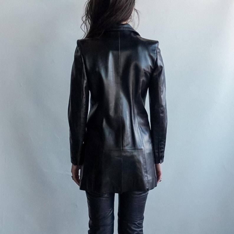 May include: A black leather blazer with a notched collar and long sleeves. The blazer has a tailored fit and a knee-length hemline. The person is wearing black leather pants. The background is a plain, light gray wall.