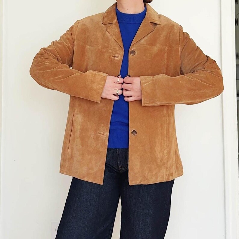 May include: A tan suede blazer with a classic collar and button closure. The jacket has long sleeves and a tailored fit. The person in the image is wearing a blue sweater and dark blue jeans.