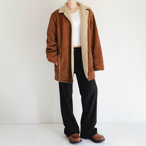 May include: A brown suede jacket with a cream-colored shearling lining and collar. The jacket has a button-up front, side pockets, and a relaxed fit. The model is wearing black pants and brown shoes.