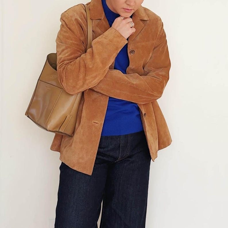 May include: A tan suede blazer with a classic collar and button closure. The blazer is paired with a blue turtleneck, dark wash denim jeans, and a tan leather tote bag with a shoulder strap.