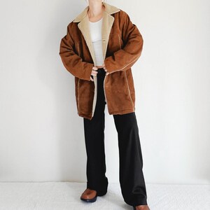 May include: A brown suede coat with a cream-colored shearling lining and trim. The coat is open, revealing a white top and black pants. The coat has a classic, vintage style.