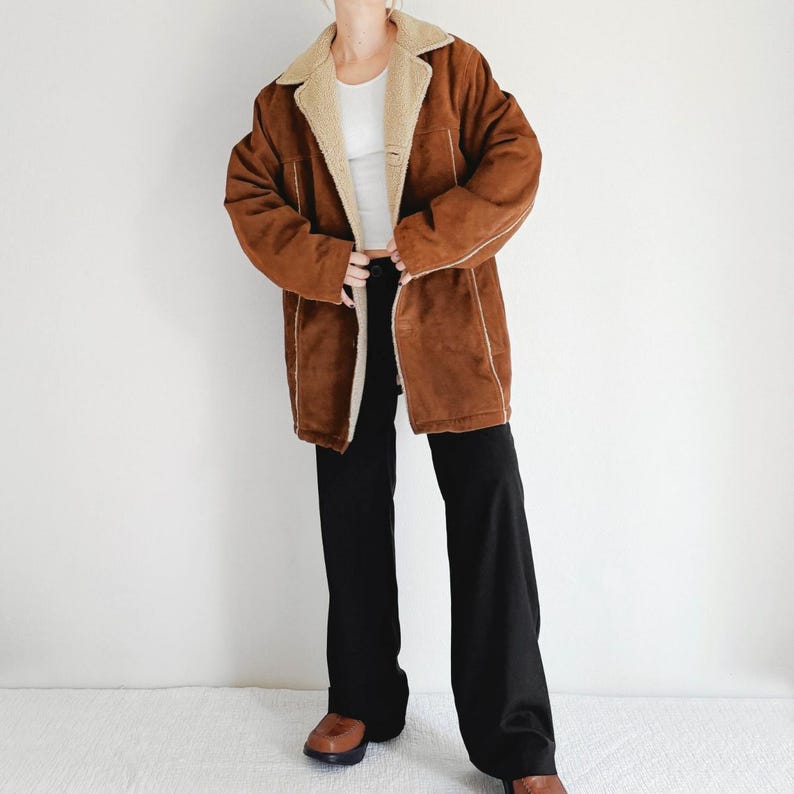 May include: A brown suede coat with a cream-colored shearling lining and trim. The coat is open, revealing a white top and black pants. The coat has a classic, vintage style.