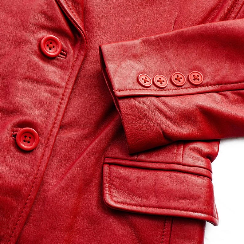 May include: Close-up of a red leather blazer. The blazer features two buttons on the front and four on the sleeve cuff. A pocket is visible on the lower right side. The leather has a smooth texture.