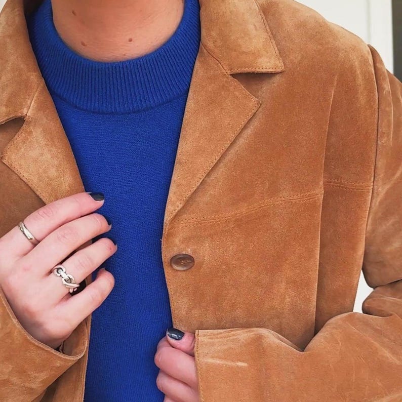 May include: A tan suede blazer with a button closure, worn over a blue sweater. The blazer has a classic collar and a tailored fit. The person is wearing silver rings.