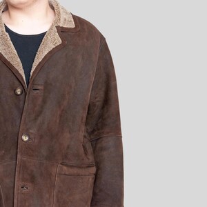 K&ouml;nnte beinhalten: Ein brauner Wildledermantel mit Schafsfellkragen und -futter. Der Mantel hat eine Knopfleiste, zwei Vordertaschen und ein klassisches Design. Ein warmes Oberbekleidungsst&uuml;ck.