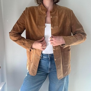 May include: A tan suede jacket with a mandarin collar and button closures. The jacket has two front pockets and is worn over a white top and blue jeans. The jacket's color is a warm, earthy tone.
