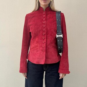 May include: A red suede jacket with a mandarin collar and button-up front. The jacket has long sleeves and small pockets. A black Dior shoulder bag with a silver buckle is worn over the shoulder. The model is wearing dark blue jeans.