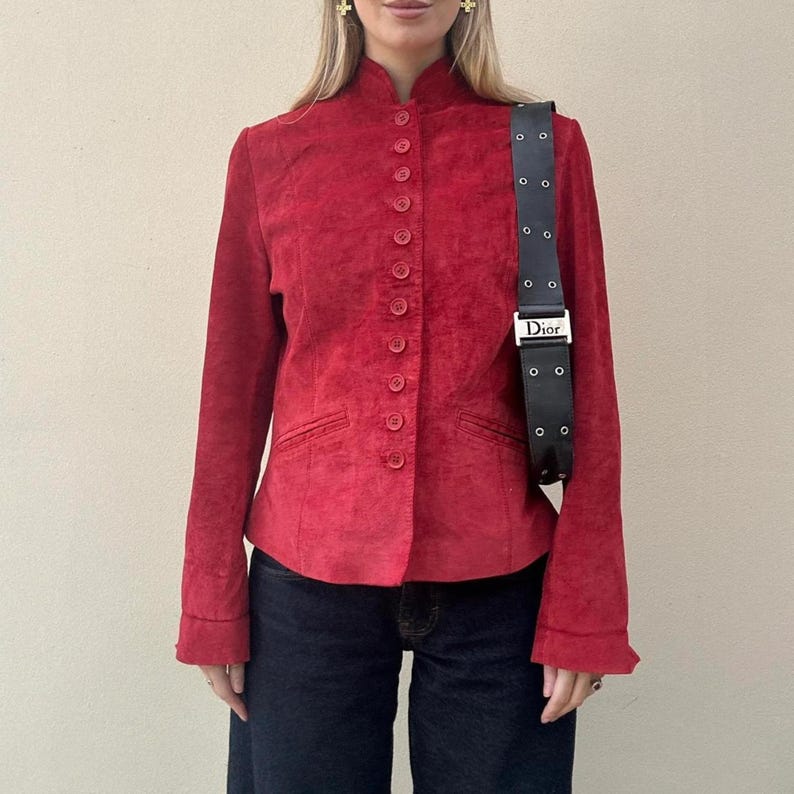 May include: A red suede jacket with a mandarin collar and button-up front. The jacket has long sleeves and small pockets. A black Dior shoulder bag with a silver buckle is worn over the shoulder. The model is wearing dark blue jeans.