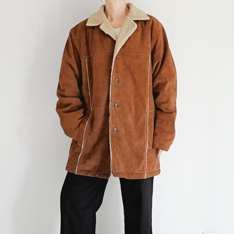May include: A brown suede jacket with a cream-colored shearling collar and lining. The jacket has a button-up front, two side pockets, and white trim along the seams. The jacket is worn over a white shirt and black pants.