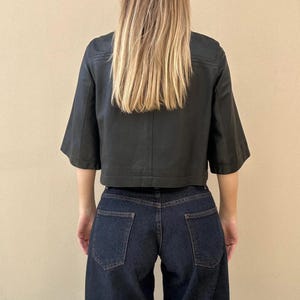May include: A black cropped jacket with short, wide sleeves and a boxy fit. The jacket is paired with dark wash denim jeans. The model's back is facing the camera, with long blonde hair.