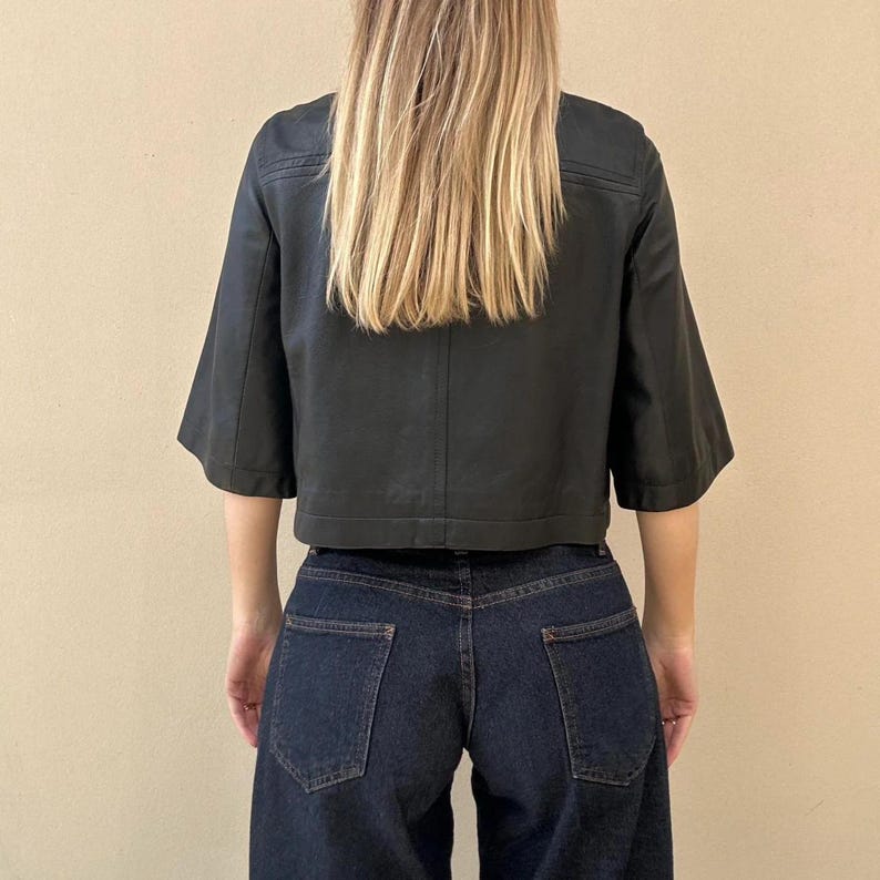 May include: A black cropped jacket with short, wide sleeves and a boxy fit. The jacket is paired with dark wash denim jeans. The model's back is facing the camera, with long blonde hair.