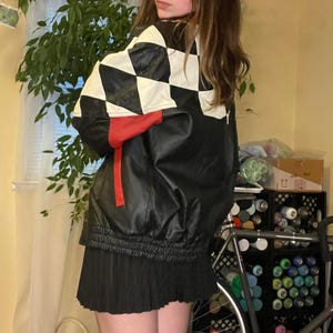 Puede incluir: Chaqueta de cuero negra con un diseño a cuadros blanco y negro en la parte superior de la espalda y los hombros. La chaqueta tiene un acento rojo en la manga y una falda plisada negra.